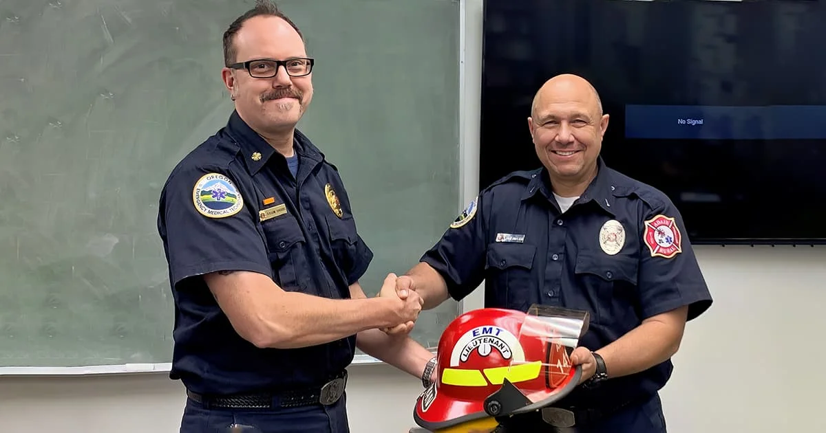 Two emergency service workers in dark blue uniforms shaking hands, with a red firefighter helmet in the foreground.