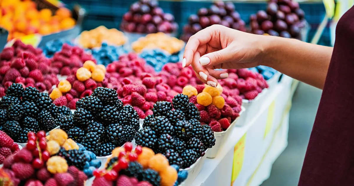 A hand selecting berries from a display of colorful fresh berries including blackberries, raspberries, and golden berries.