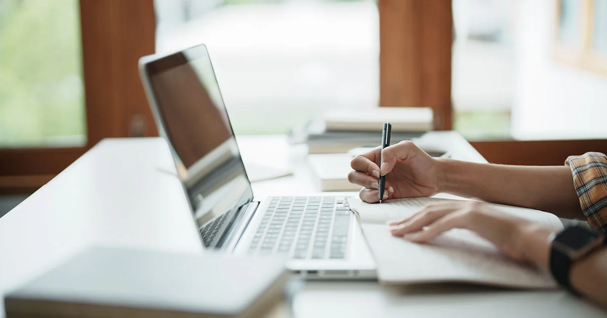 A laptop and a hand writing on a piece of paper at a desk near a window.