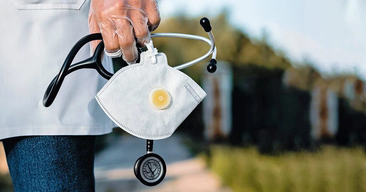 Medical professional holding a protective mask and stethoscope, with blurred outdoor background.