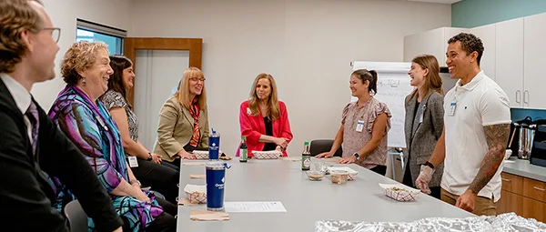 Group of people standing around a conference table in an office meeting room, with drinks and food containers visible.