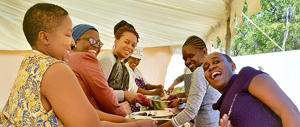 Group of women preparing food together in a white tent, smiling and laughing while working with metal trays and an orange mesh bag.