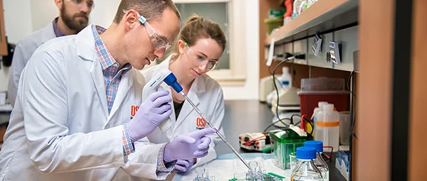 Two researchers in white lab coats and protective glasses working together in a scientific laboratory, using pipettes and laboratory equipment.