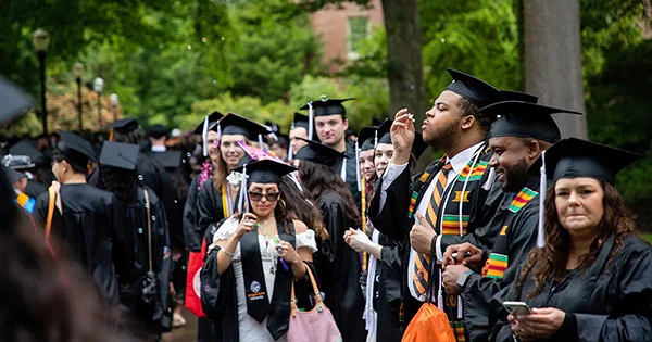 Group of graduates in black caps and gowns standing together outdoors, some wearing colorful stoles, with trees and a building in the background.