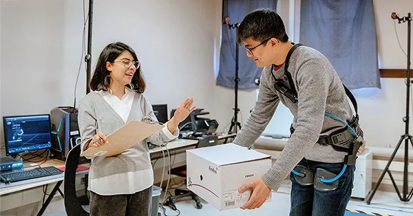 Two people in a technical workspace, with computer monitors and equipment, interacting near a white box.