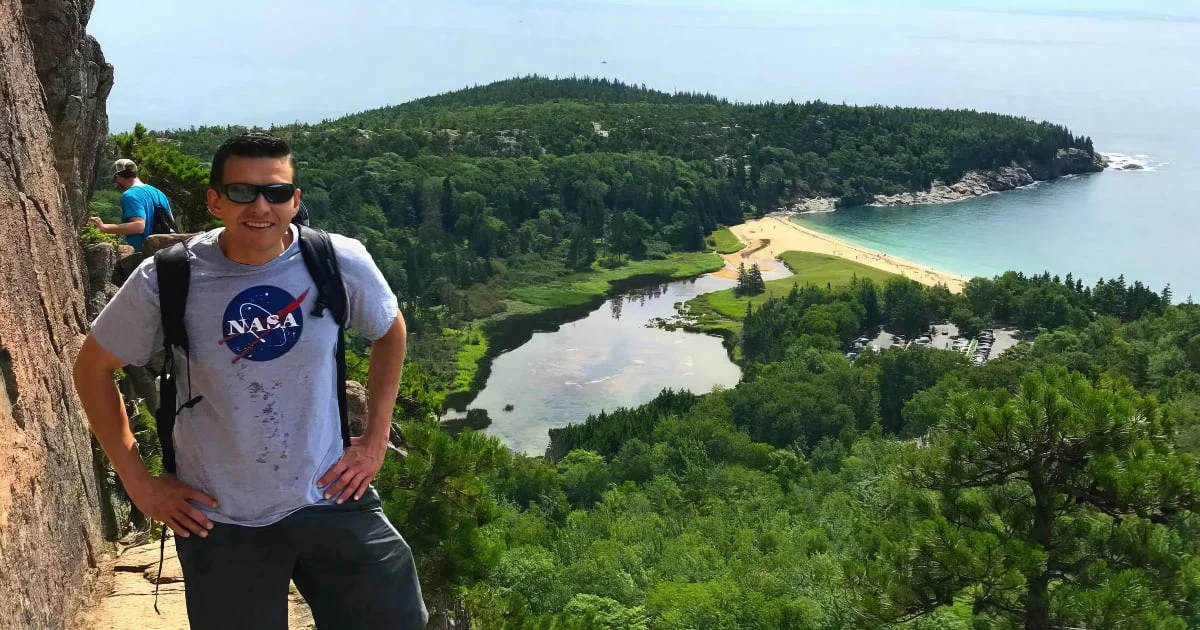Andres wearing a NASA t-shirt standing on a rocky cliff overlooking a scenic coastal landscape with a beach, forest, and lake.