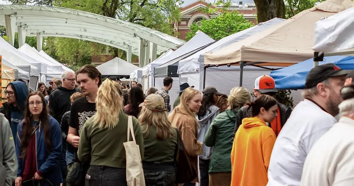 Outdoor market with white tents and many people walking around, with trees and an arched structure in the background.