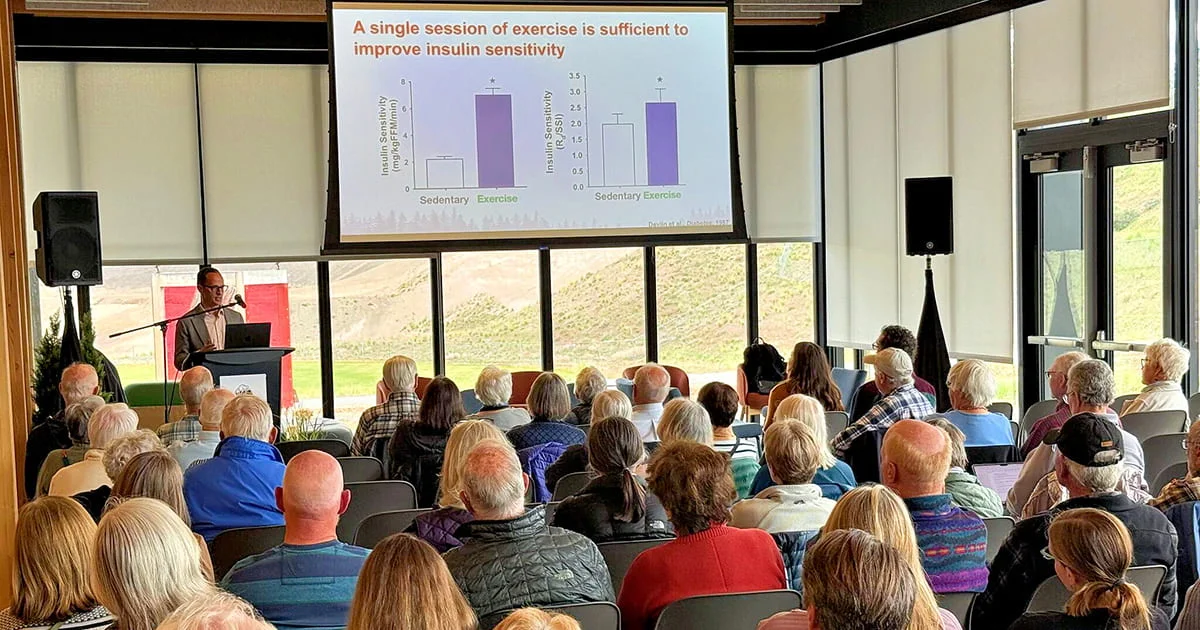 A presentation about exercise and insulin sensitivity with a speaker at a podium and an audience of mostly older adults seated in a room with large windows.