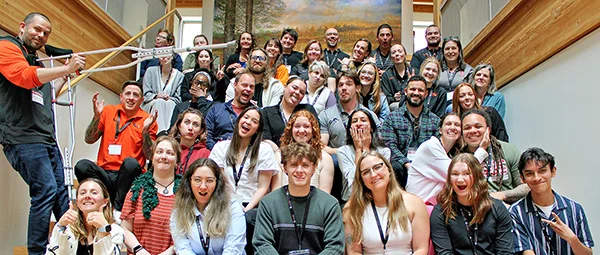 Large group photo of people sitting and standing on wooden stairs, with a landscape painting in the background, wearing lanyards and casual clothing.