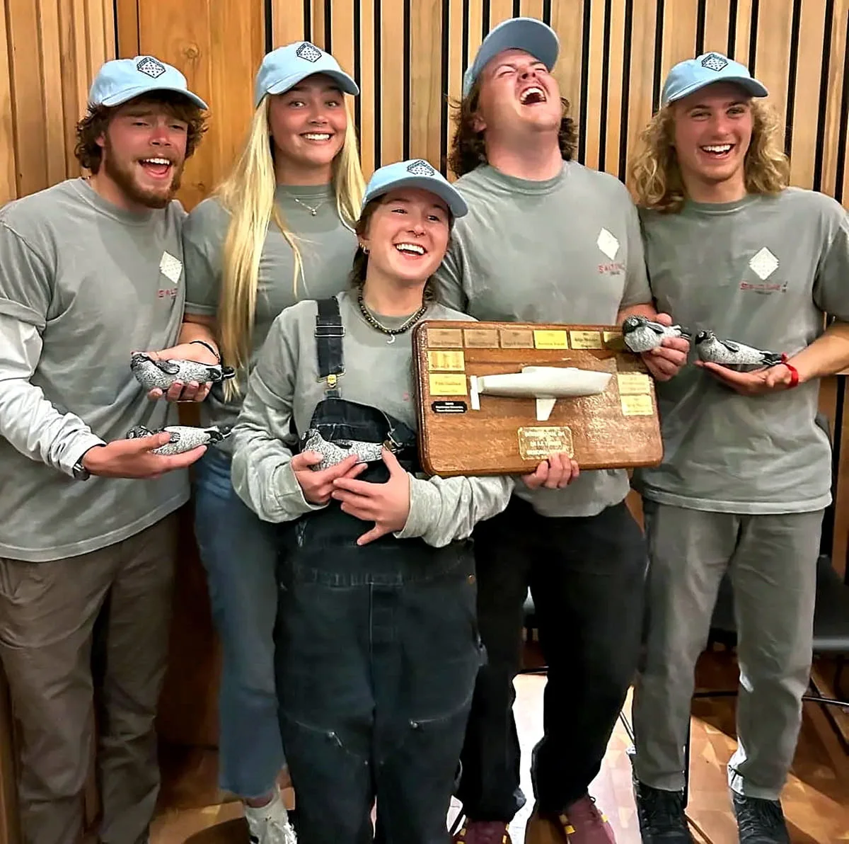 Five young people in matching gray shirts and blue caps, holding a wooden plaque, smiling and laughing together.