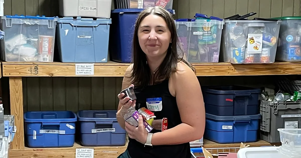 A person standing in a storage area filled with bins and boxes, holding several items in their hands. The person has long hair and is wearing a black tank top. The background features shelves with various containers and labels.