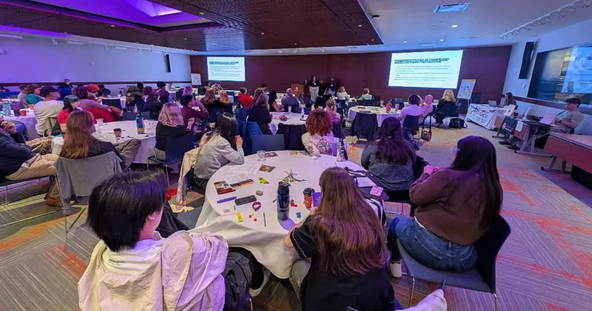 A conference or workshop setting with a large audience seated at round tables. The room is well-lit, and there are presentations displayed on screens. Attendees are engaged, some taking notes or using devices.