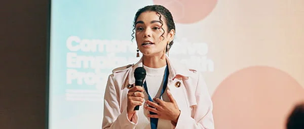 A woman speaking at a conference or event, holding a microphone. She is wearing a light pink jacket and a white top, with a lanyard around her neck.