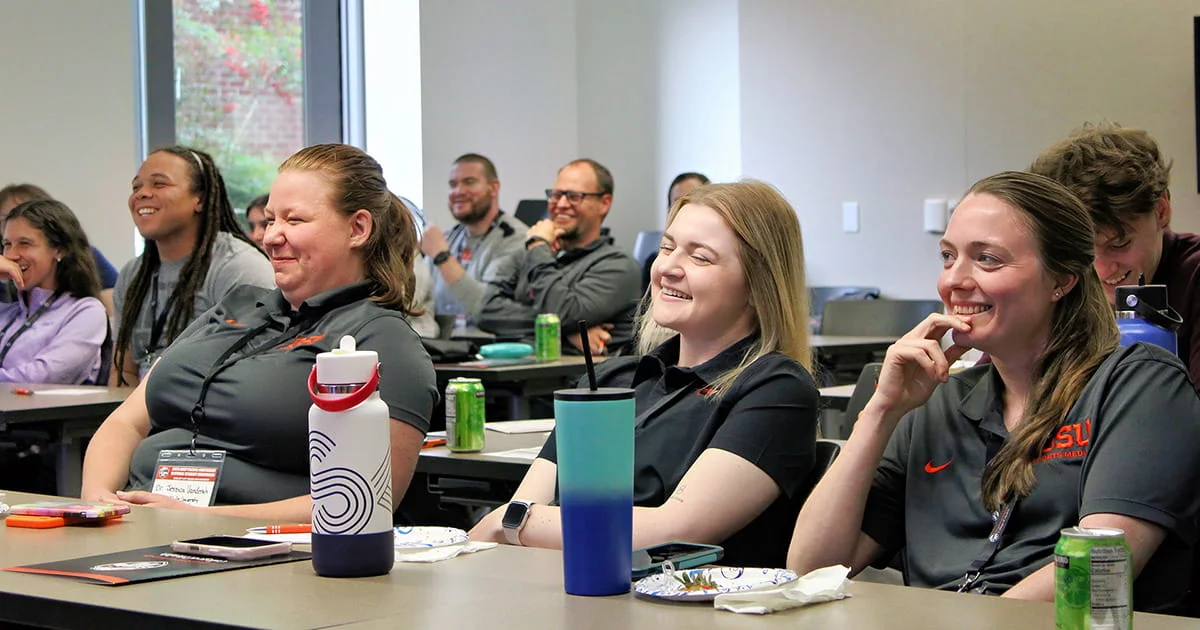 A group of people sitting in a classroom or conference room, smiling and engaged in a presentation or discussion.