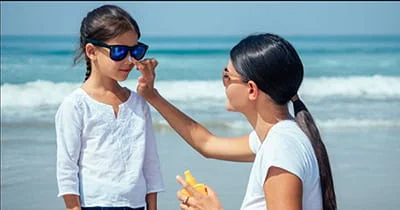 A woman applies sunscreen to a young girl's nose on a sunny beach, with the ocean in the background.