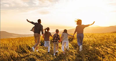 Group of people walking through a golden wheat field at sunset, with mountains in the background, viewed from behind.