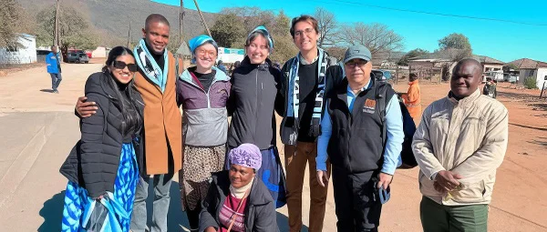 A group of seven diverse individuals, some wearing scarves, standing outdoors in an arid African village setting with mountains in the background.