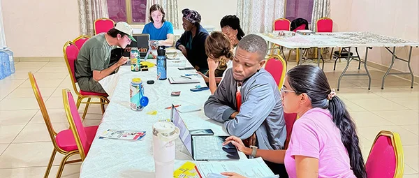Several people collaborating on computers and documents around a large table during a workshop or meeting.