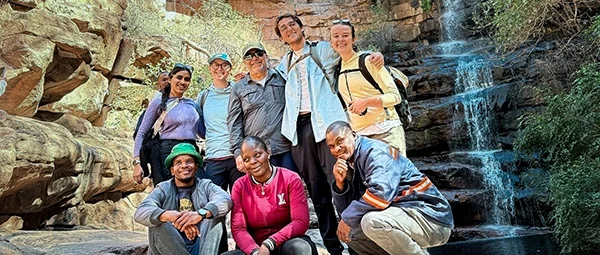 A joyful group of eight, including five standing and three crouching in the foreground, with cascading water and weathered rock cliffs behind them.