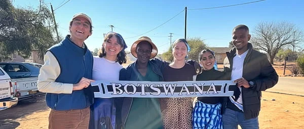 Six people, including men and women of various ethnicities, are gathered outdoors under a clear sky, standing on a dirt road next to some vehicles and trees, proudly displaying a long, narrow banner with the word "BOTSWANA" written across it.