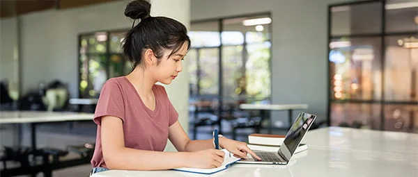 A student engaged in focused study, using a laptop and a notebook in what appears to be a university common area or library.