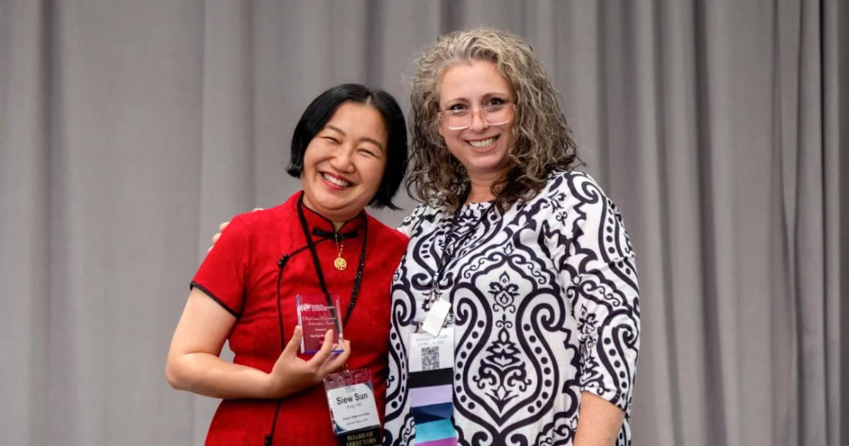 A woman in a red outfit holds an award trophy and smiles next to another smiling woman with curly hair and a patterned shirt, likely at an event or conference