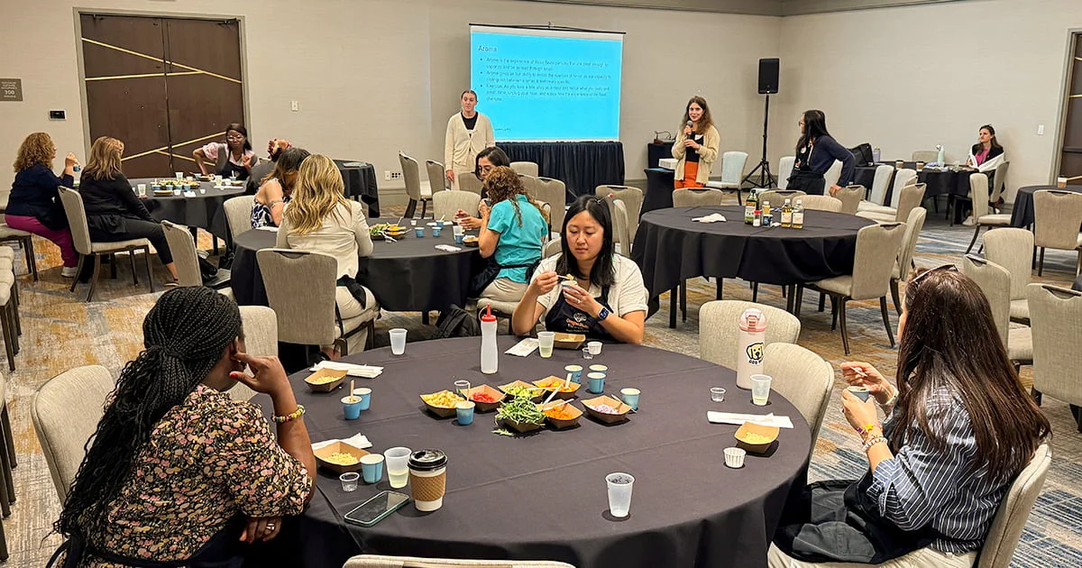 Attendees participating in a workshop or conference session, with many individuals at tables engaged in a hands-on activity involving food, and a speaker presenting at the front of the room.