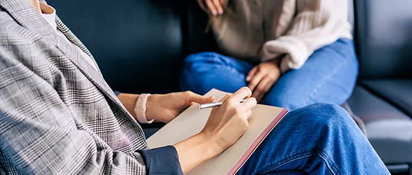 A counselor or behavioral health professional takes notes during a session, focusing on active listening and documentation, with a client partially visible.
