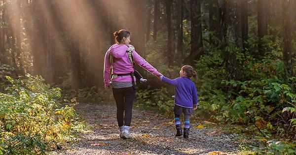 A caregiver and child walking hand in hand on a forest path with sunlight filtering through trees.