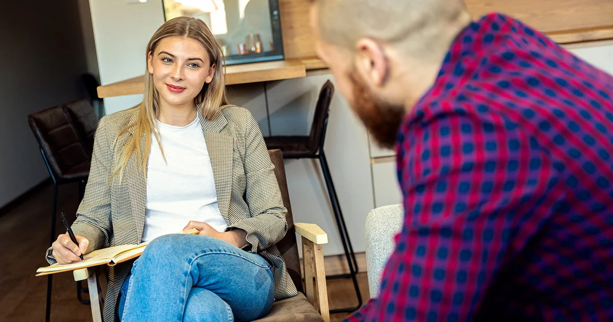 A female counselor or therapist takes notes during a session, engaging with a client (partially seen), suggesting a professional behavioral health consultation