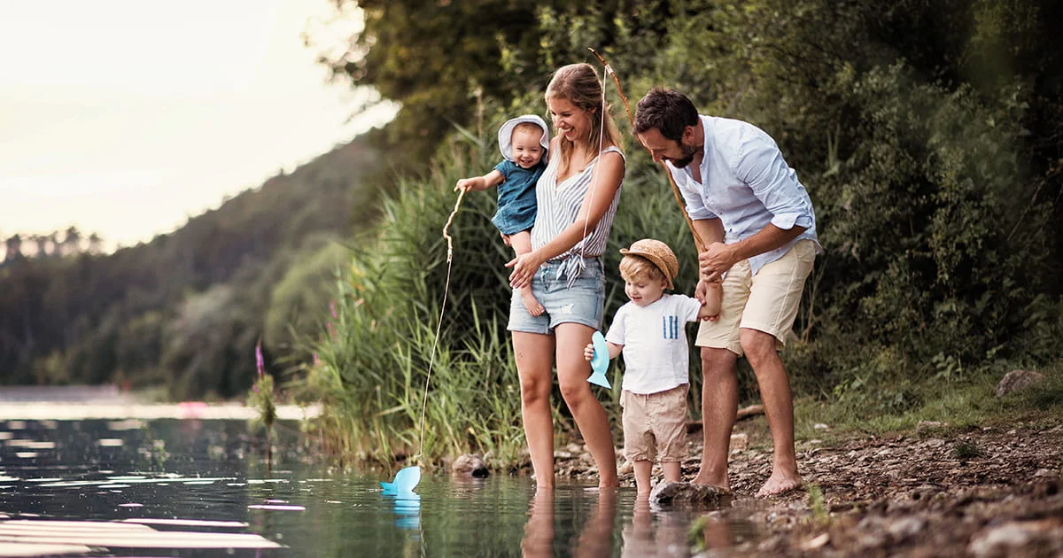 A family of four standing by a lake, playing with a blue object in the water, surrounded by greenery and trees.