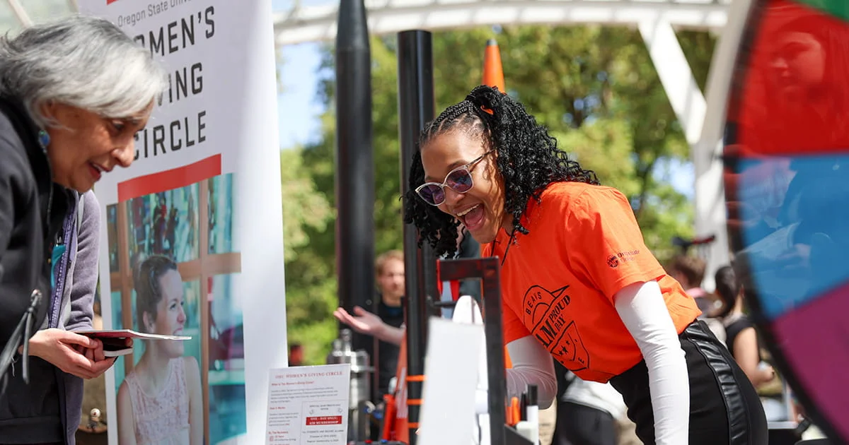 Two women at an outdoor event booth with a "Women's Giving Circle" sign, one in an orange shirt and sunglasses, the other in a gray top.