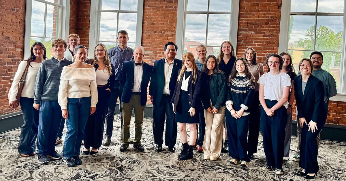 A large group of young adults and a few older adults are assembled for a photograph in an interior space characterized by tall windows, red brick walls, and a patterned carpet, radiating a positive atmosphere.