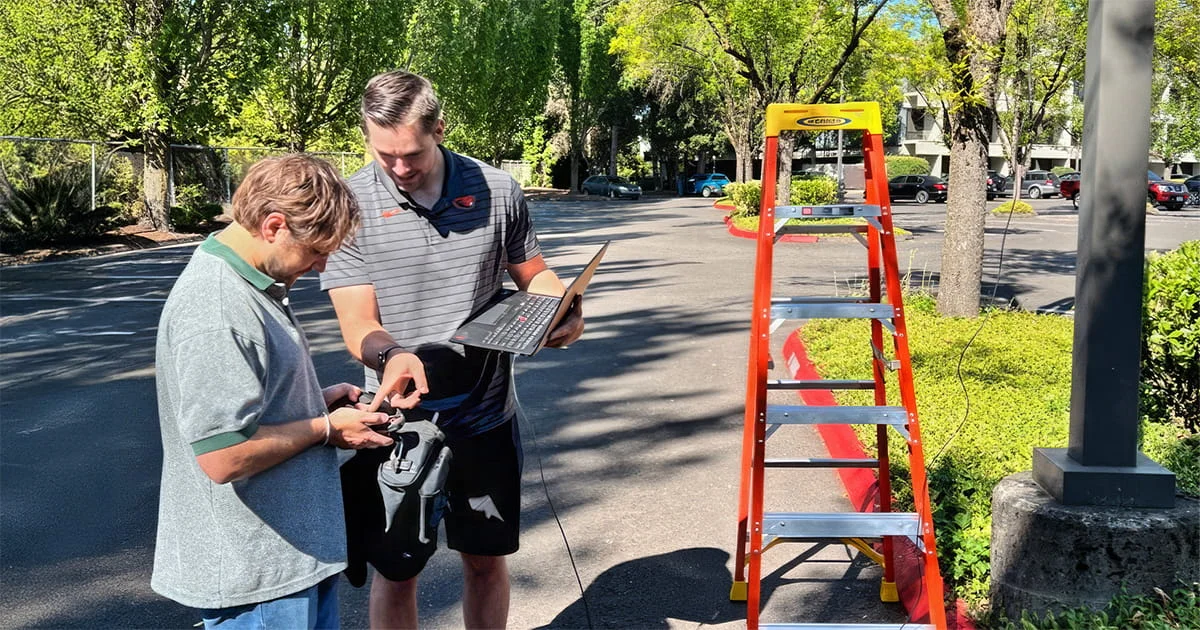 Two men collaborate to calibrate and test a sound monitoring system next to a red ladder and a black pole, outdoors.