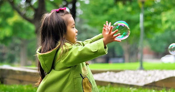 A smiling young girl with long dark hair and a pink bow, wearing a green coat, playfully reaching out to touch a large, iridescent soap bubble in a grassy park.