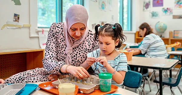 A woman in a pink patterned hijab and dress leans over a young girl with pigtails to help her work on a project with an egg carton. They are at a small table in a bright classroom with other children and an adult visible in the background.