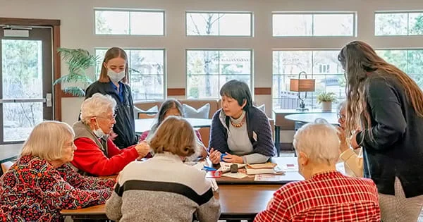 A student in a healthcare setting is assisting a group of senior citizens during an activity at a table.