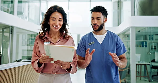 Two healthcare professionals standing in a modern medical facility, one in a pink blouse holding a tablet, the other in blue medical scrubs gesturing while talking.
