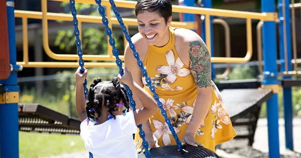 A woman smiles as she pushes a child on a swing at a playground.