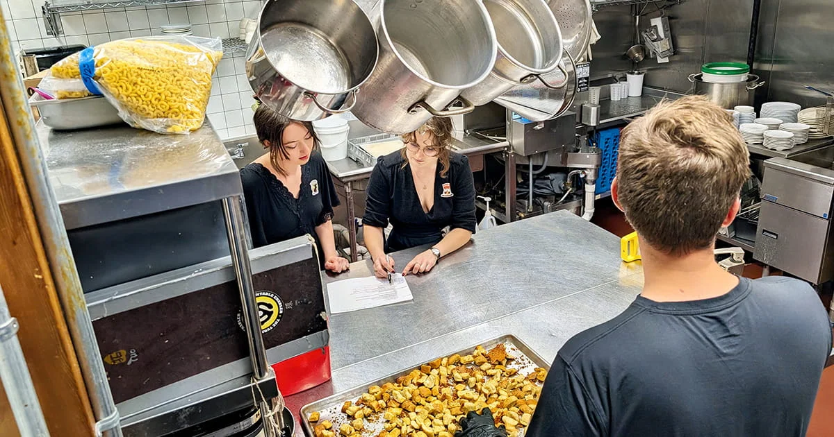 A busy commercial kitchen with three staff members working; two are seated discussing paperwork near hanging stockpots.