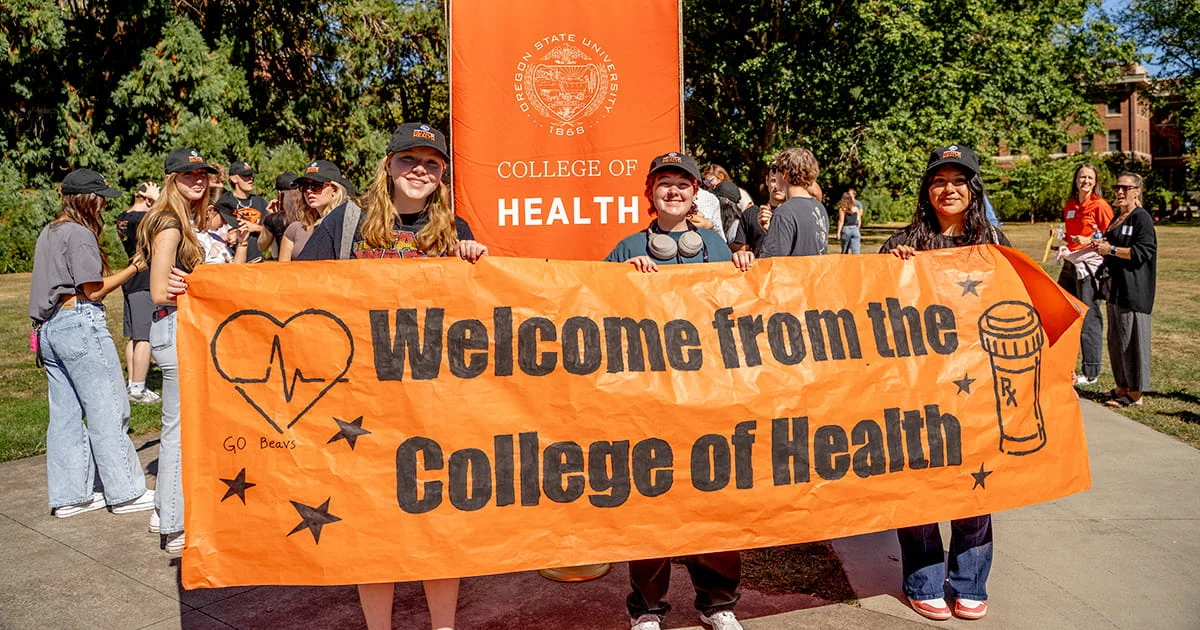 Students at Oregon State University College of Health hold a welcome banner on a sunny day during an outdoor campus event, with a large school banner visible behind them.