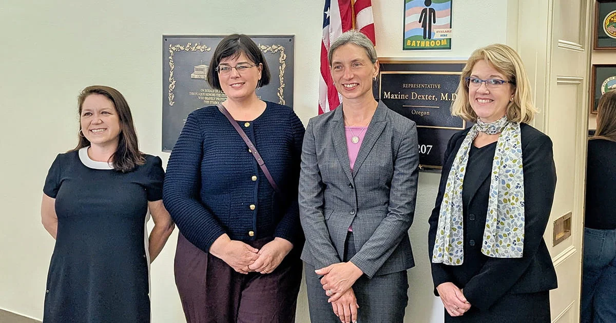 Four women standing in a well-lit hallway with a plaque for Representative Maxine Dexter, M.D. (Oregon) visible, along with an American flag and a sign for an inclusive bathroom.