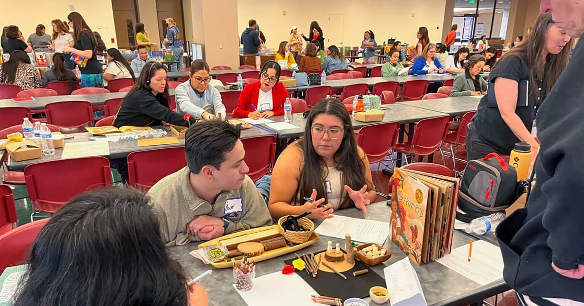 A woman gestures as she speaks to a small group seated around a table covered in papers, writing tools, and a children's book. Many other participants are visible at tables across the large room.