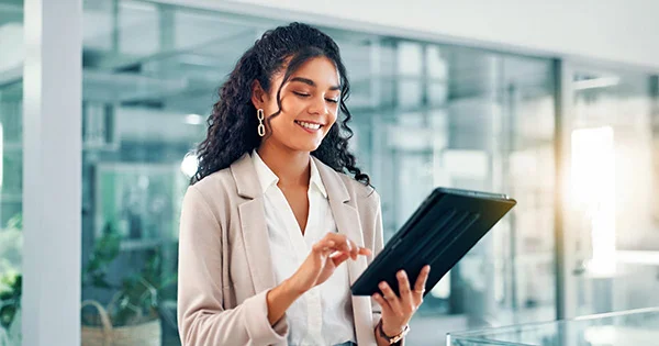 A professional woman in a light-colored blazer using a tablet in a modern glass office environment, smiling and looking down at the device.