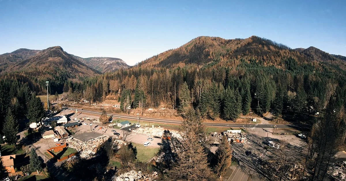 Panoramic photo of a small rural town devastated by fire. The foreground shows flattened structures and scattered debris along a main road, contrasting with the scarred mountains rising in the background.