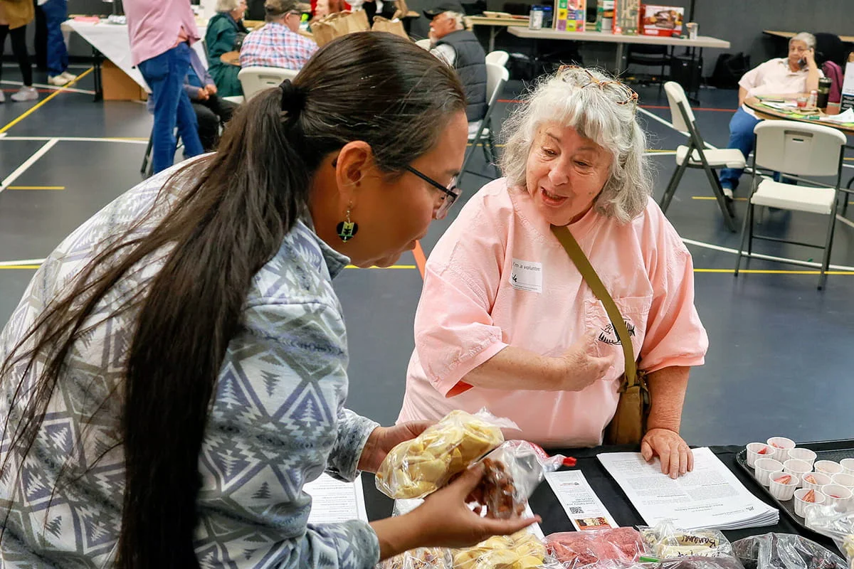 A woman with long dark hair holds up two bags of dried food to show an older attendee with white hair seated at a display table, discussing food preservation.