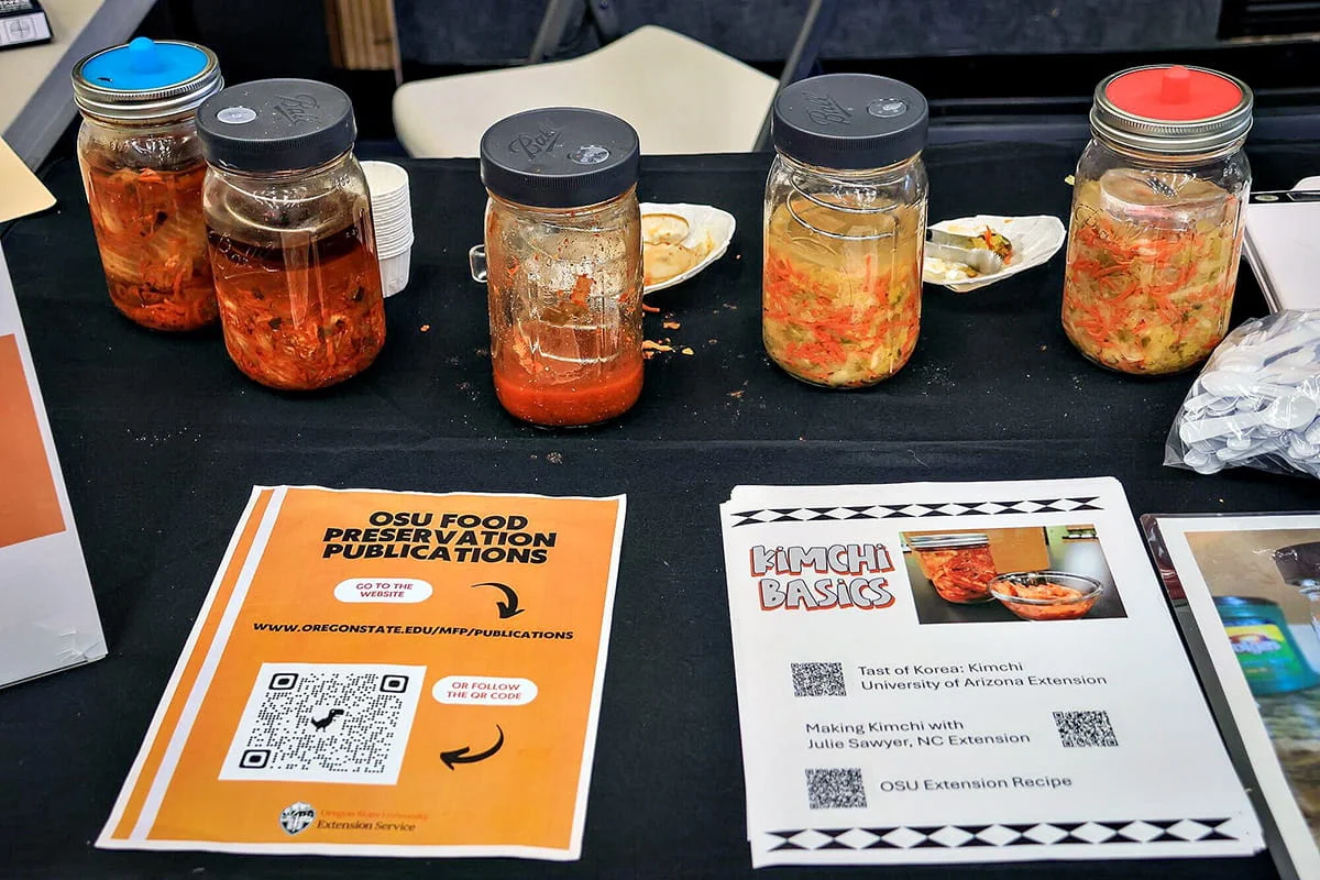 Four glass jars of homemade kimchi and other preserved foods displayed on a black table next to informational handouts about kimchi and food preservation publications.