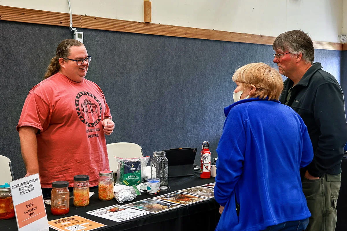 A male instructor in a red shirt talks to an older couple at a table display featuring jars of sauerkraut, a demo schedule, and informational pamphlets about food preservation.