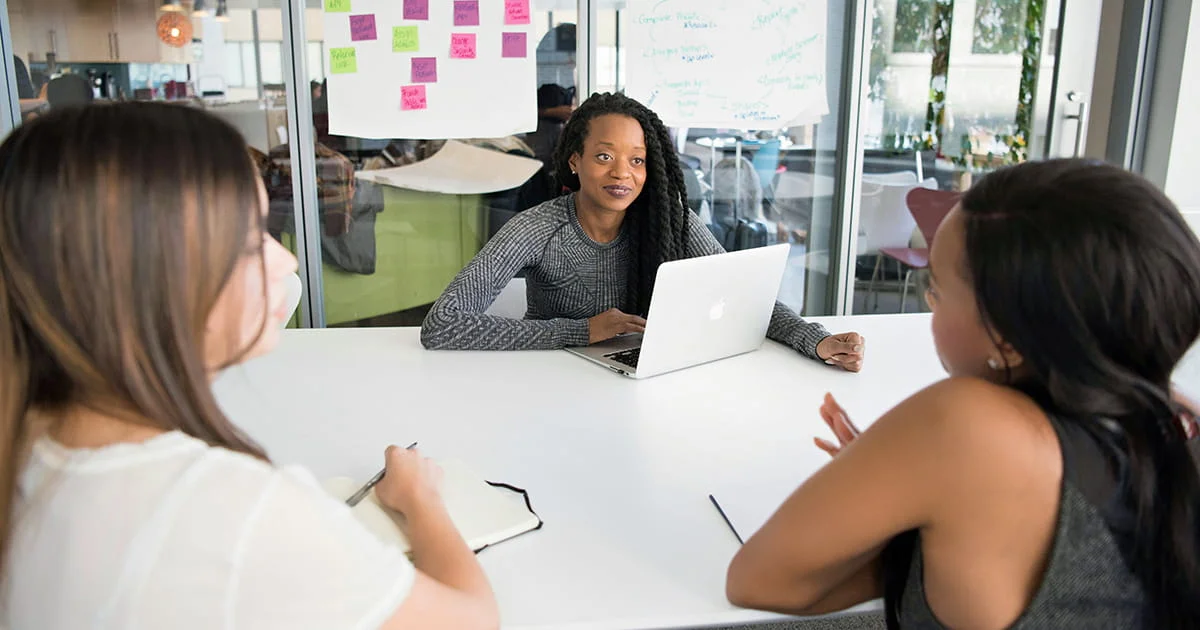 A woman seated at the center of a white table is speaking to two colleagues. A laptop is open in front of her, and post-it notes are visible on a glass wall in the background.
