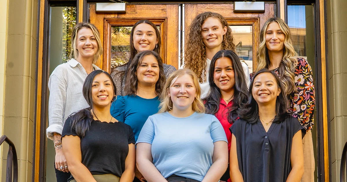 Group portrait of nine smiling, diverse young women, likely the OSUDI 2026 cohort, posing on steps in front of a large wooden door.